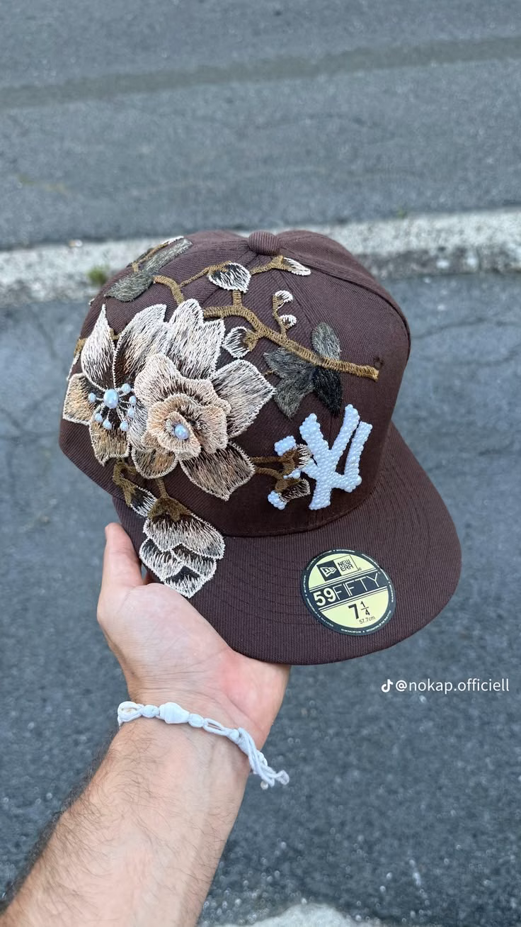 A hand holds a brown New York Yankees fitted cap with large floral appliqués.
