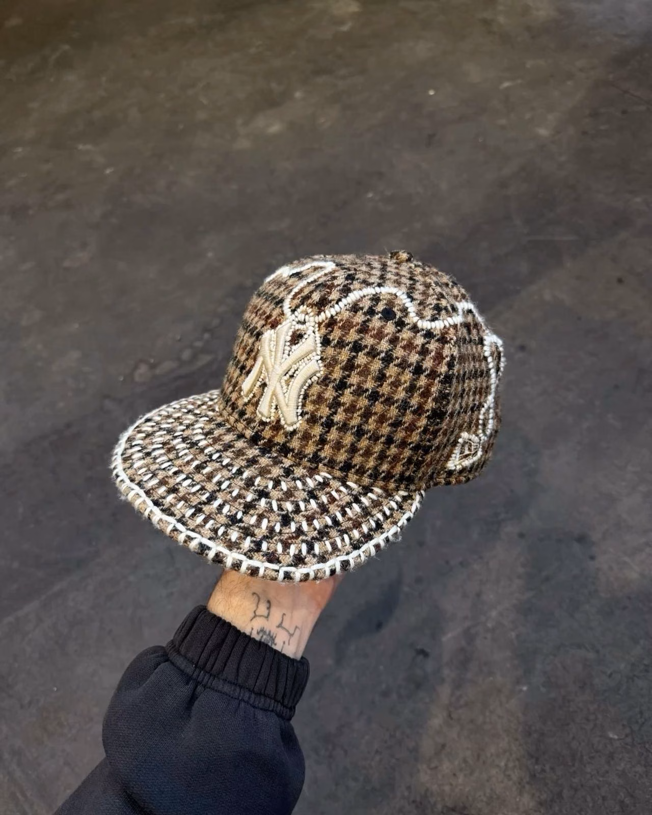 A hand holds a brown checked New York Yankees fitted cap with fringed details.