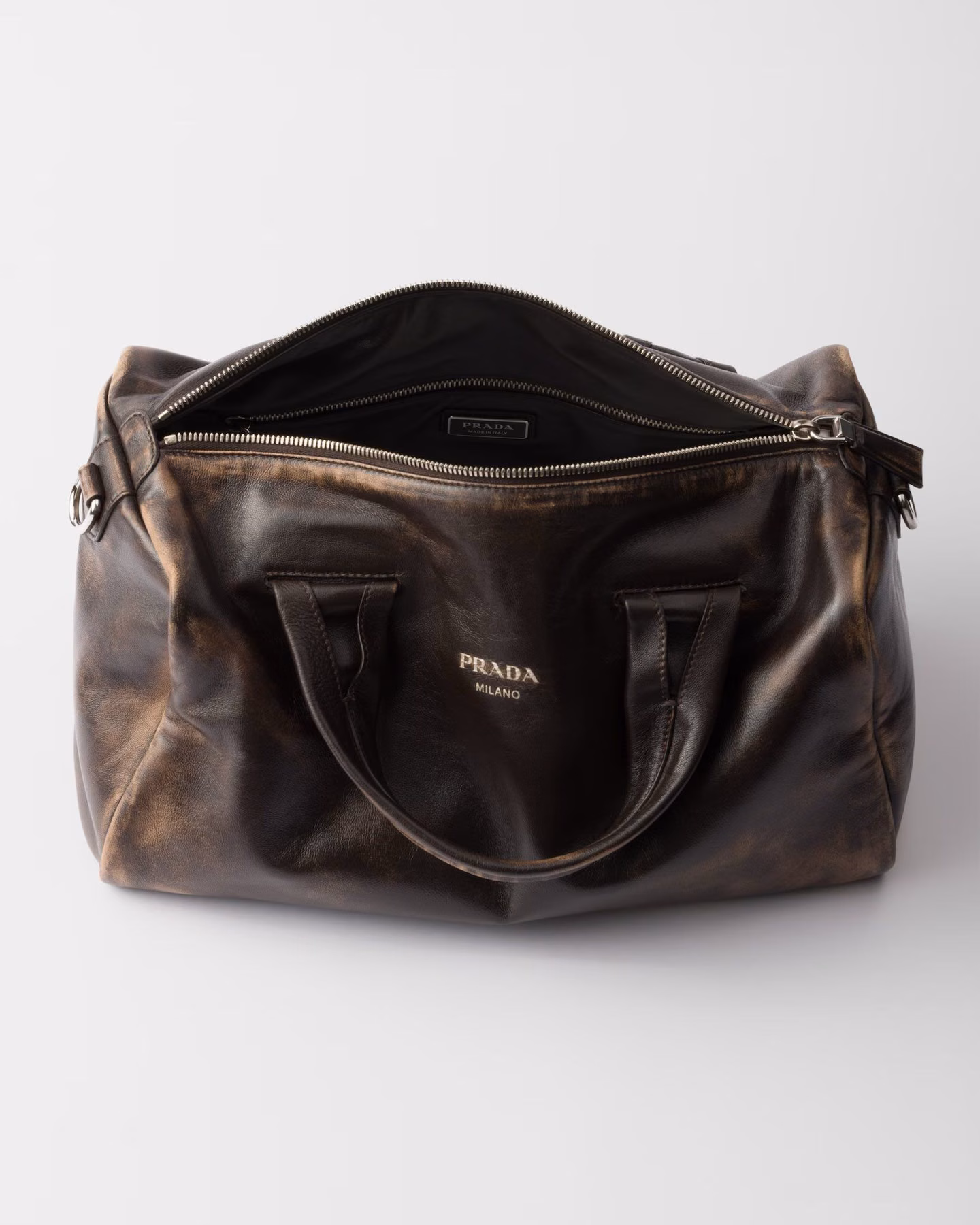 A dark-brown Prada travel bag in patinated leather against a light background.