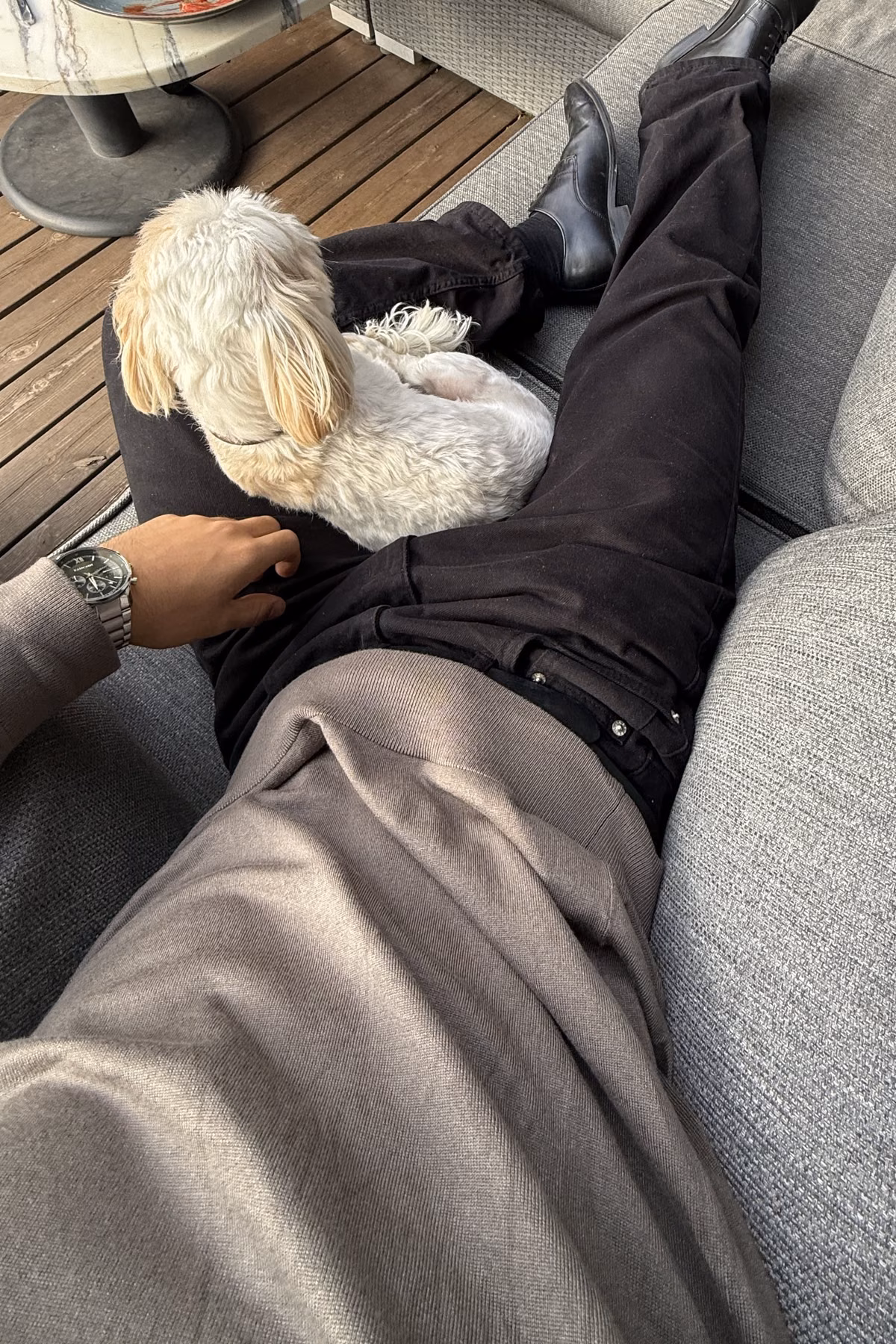 A person in a taupe knit, black jeans, black leather boots and a steel watch sits on an outdoor sofa with a small white dog asleep across their lap.