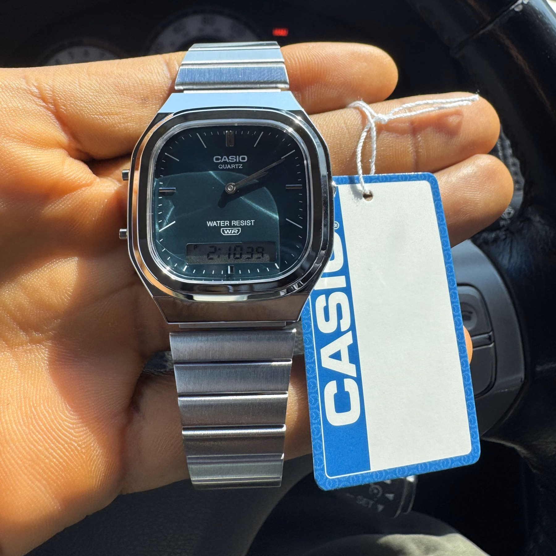 A square steel Casio digital watch with a blue tag held in one hand.