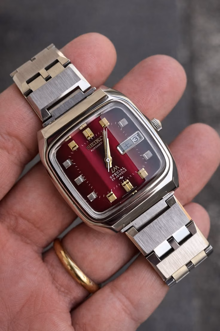 A rectangular vintage steel watch with a burgundy dial held in one hand.