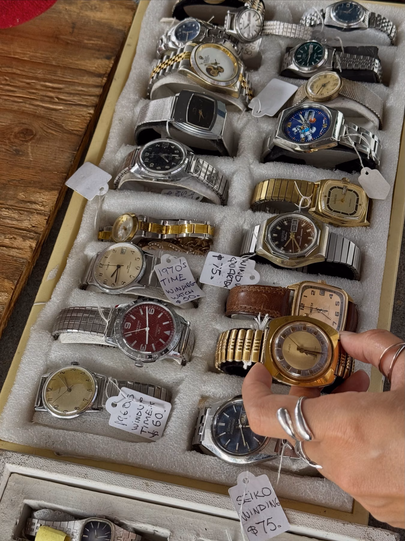 A tray filled with assorted vintage watches and handwritten price tags while a hand chooses among them.